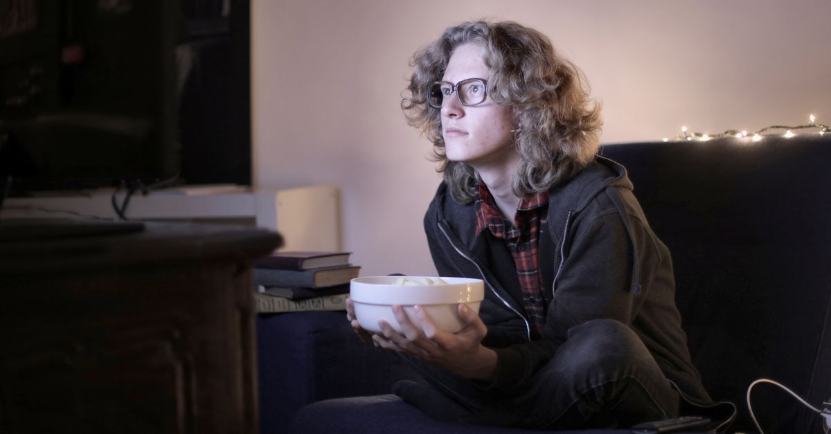 Concentrated male teenage with long hair in casual wear and eyeglasses sitting on couch with bowl of popcorn and watching interesting movie while resting in cozy living room at home